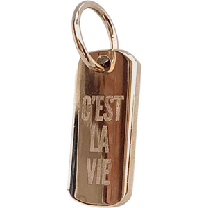 A gold-colored polished brass pendant with the phrase 'C'est la vie' engraved on it, featuring a lobster clasp fastening.