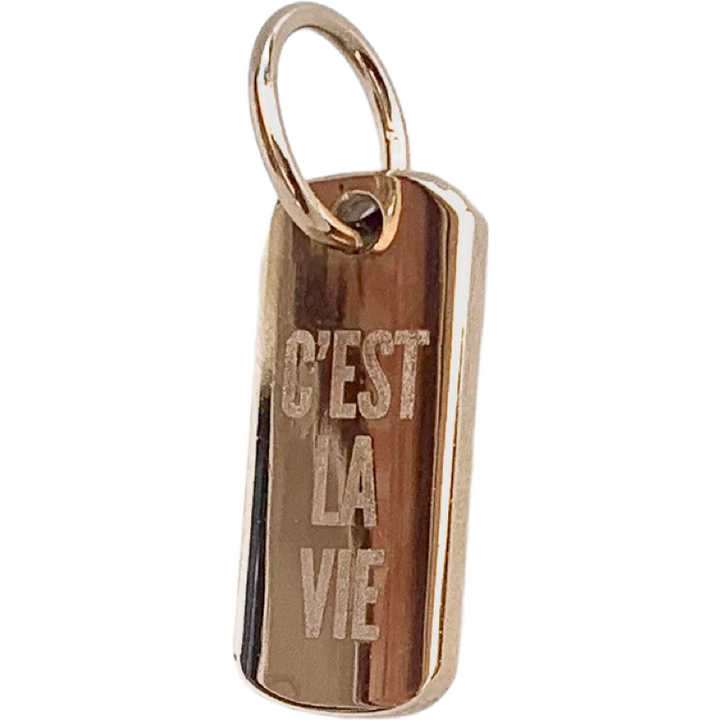 A gold-colored polished brass pendant with the phrase 'C'est la vie' engraved on it, featuring a lobster clasp fastening.